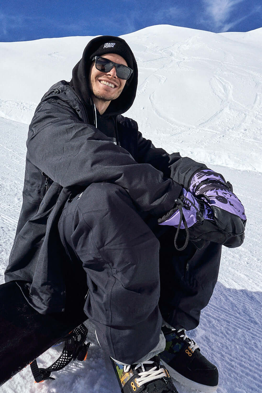 Snowboarder wearing The Team Transform x Glitterstudio Lavender mittens with Primaloft insulation, seated on a snowy slope.