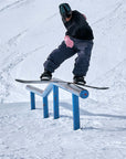 Snowboarder wearing pink gloves performing a rail slide trick on snowy terrain