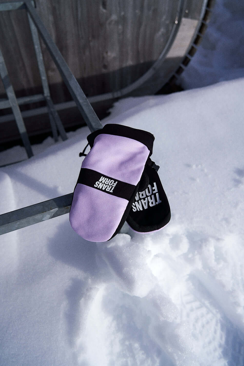 Pink and black snowboard mittens on snow with outdoor gear in the background.